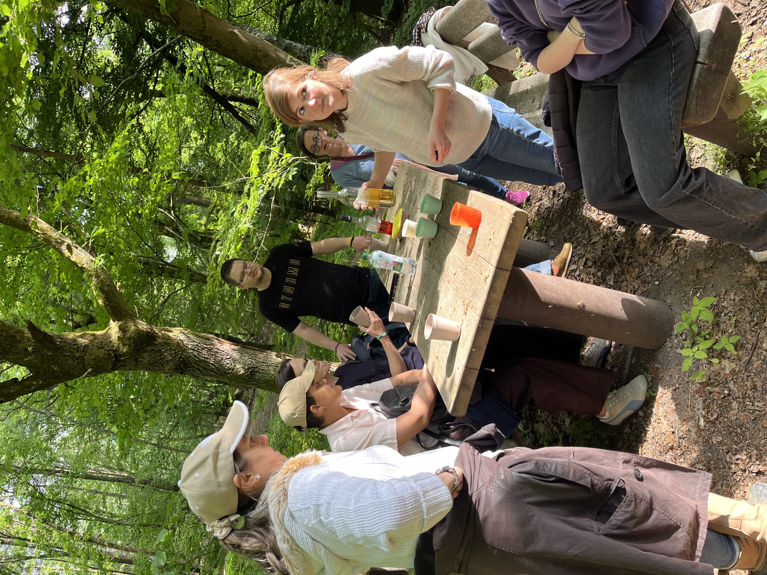 Eine Gruppe von Menschen macht eine Pause an einem rustikalen Holztisch mitten im grünen Wald. Einige sitzen oder stehen entspannt beisammen, trinken aus Bechern und unterhalten sich bei sonnigem Wetter zwischen hohen Bäumen.